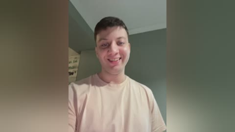 Video of a young, smiling Caucasian man with short brown hair, wearing a light beige T-shirt, standing in a room with beige walls and a white ceiling.