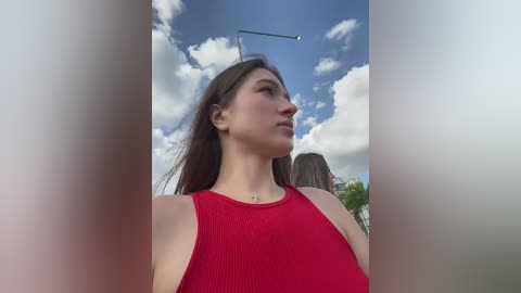 Media: Video of a young woman with long brown hair in a red sleeveless top, standing outdoors under a blue sky with scattered clouds.