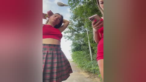 Media: Video of a young woman with light skin and short dark hair in a red crop top and plaid skirt, taking a selfie outdoors on a tree-lined path.