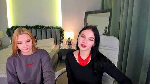 Video of two young women with fair skin and long hair, wearing casual outfits, in a modern bedroom with a bed, nightstand, and mirror.