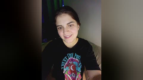 Video of a smiling young woman with fair skin, dark hair, and brown eyes, wearing a black T-shirt with a colorful, psychedelic design. She is seated indoors on a beige couch.