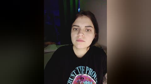 Video of a young woman with light skin, dark hair, and a black shirt featuring a colorful \"The Doors\" logo. She gazes into the camera, indoors with dim lighting and a blurred background.