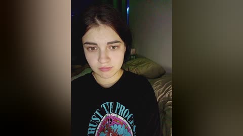 Video of a young woman with dark hair, fair skin, and a serious expression, wearing a black t-shirt with a colorful logo, in a dimly lit bedroom with green bedding and a nightstand.