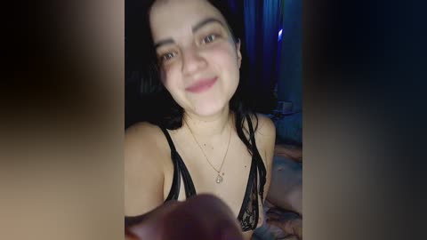 A young woman with fair skin, dark hair, and a warm smile, wearing a black lace bra, videoed indoors in a dimly lit room.