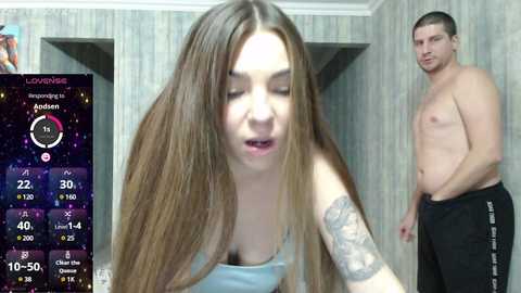 Video of a slender, fair-skinned woman with long brown hair, wearing a light blue tank top, looking seductively towards a shirtless, muscular man in black sweatpants in a dimly-lit room.