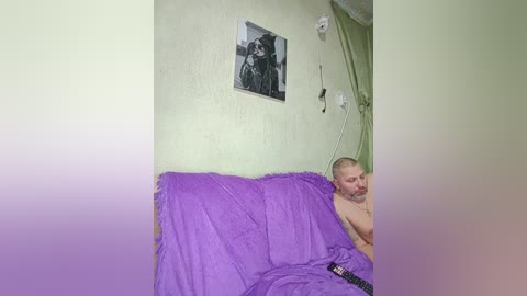 A video shows a bearded, shirtless man in a purple bedspread, lying on a green couch, with a black-and-white framed photo and electrical outlets on the wall behind him.