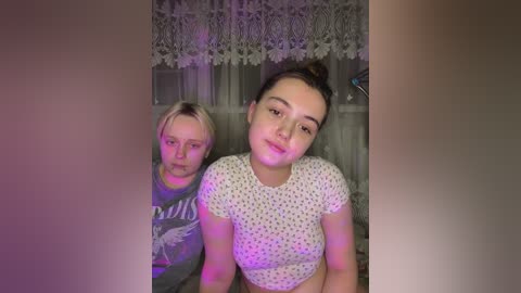 Media: Video of two young girls with fair skin and light brown hair, one in a grey T-shirt, the other in a white polka-dotted top. They stand in a room with lace curtains and purple lighting, creating a cozy, intimate atmosphere.