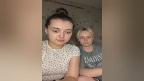 Media: Video of two young women in casual clothes; one with dark hair in a bun, another with blonde hair, both looking serious, in a dimly lit room with white walls.