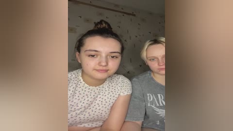 Media: Video of two young women, one with light skin and dark hair in a bun, wearing a floral top, and another with blonde hair, gray t-shirt with \"PARIS\" text. They are indoors, partially obscured by a wooden frame.