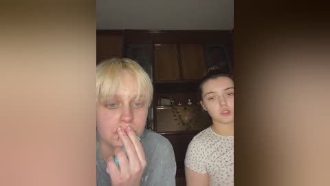 Media: A video shows a blonde woman with fair skin, wearing a gray shirt, holding her finger to her mouth, and a brunette woman in a white polka-dot top, both indoors with wooden cabinets in the background.