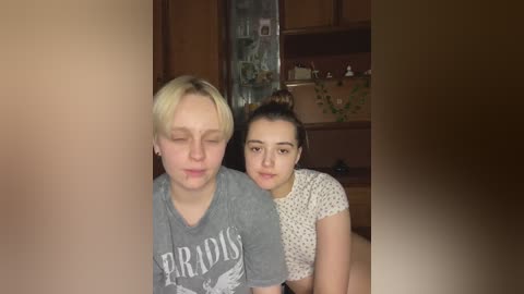 Media: Video of two young women, one with short blonde hair in a gray T-shirt, the other with dark hair in a white polka-dotted top, in a cozy, wooden-paneled room with shelves and green plants.