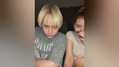 Media: Video of two young women with short blonde hair, one in a gray \"Paradise\" t-shirt, the other in a white polka dot top. They have a serious expression, indoors with dark wooden background.