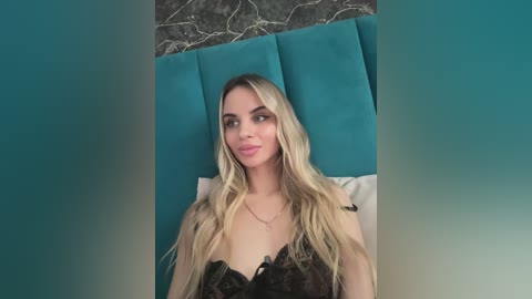 Media: Video of a blonde woman with long, wavy hair, wearing a black lace bralette, sitting on a teal upholstered headboard with white pillows in a bedroom.