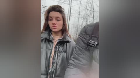 Media: Video of a young Caucasian woman with long brown hair, wearing a black quilted jacket, walking outdoors in a forest setting with bare trees in the background.