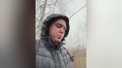 Media: Video of a pale-skinned man with short, dark hair, wearing a gray puffy jacket and hood, looking down thoughtfully, surrounded by bare trees and overcast sky.