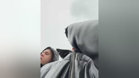 Media: Video of a young person, possibly male, with light skin, sleeping on a gray quilted blanket against a plain white wall. The image is taken from a low angle, giving a distorted, abstract perspective.