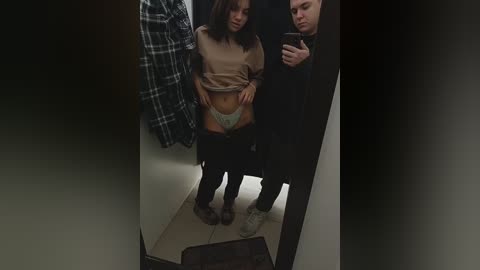 Media: A video of a young woman in a beige top, black pants, and green underwear, standing in a changing room with a man holding a phone, taking a mirror selfie.