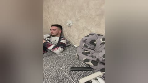 Media: Video of a young man in a cramped, dimly lit room with peeling wallpaper, wearing a patterned sweater, sitting on a leopard print carpet, surrounded by disorganized blankets and cables.