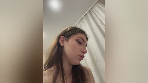 Media: A video of a young woman with long brown hair and striking red eye makeup, wearing a beige top, taken indoors with a beige curtain in the background.