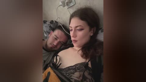 Media: Video of a woman with curly brown hair, wearing a black lace top, lying next to a man in a dark jacket, both asleep on a gray pillow in a dimly lit room.