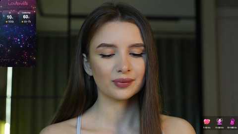 Media: Video of a young woman with long, straight brown hair, wearing minimal makeup and a light gray top. She has a serene expression, set against a blurred indoor background.