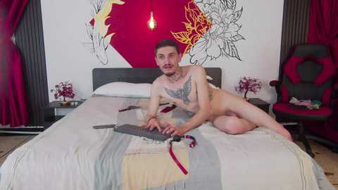 Video of a nude, fair-skinned, tattooed man with short hair lying on a bed with a red and black floral backdrop, holding a sword, in a modern bedroom with red curtains and a gaming chair.