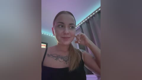 Media: Video of a light-skinned woman with long blonde hair, wearing a black tank top, showing a detailed tattoo on her chest. She's standing in a room with a purple LED strip light and a window with grey curtains.