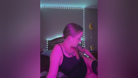 Media: A video of a young woman with light skin and blonde hair, wearing a black camisole, sitting on a bed with a wooden headboard, bathed in pink and purple light.