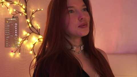 Video of a young woman with long red hair, wearing a black choker with a heart pendant, in a softly lit room with warm, ambient lighting and fairy lights.