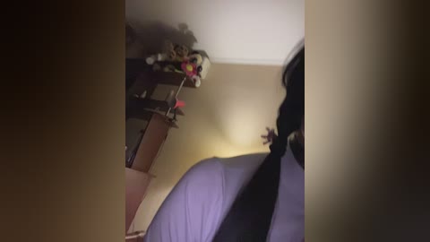 A video of a dimly lit bedroom corner showing a bed with white sheets, a shelf with toys, and a partially visible person in black clothing standing in the background.
