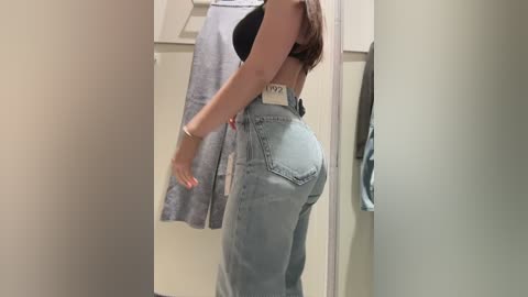 Video of a woman in a fitting room, wearing a black bra and high-waisted light blue jeans. Her back is to the camera, highlighting her curvy figure and buttocks.