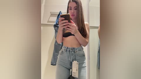 Video of a young woman with long brown hair, holding a smartphone, taking a mirror selfie in a dressing room. She wears a black bra and high-waisted light blue jeans. A denim jacket hangs behind her.