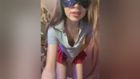 Media: Video of a young, light-skinned woman with long, straight brown hair, wearing a gray shirt and red skirt, black blindfold, and a blue and red mask, leaning forward, indoors with a cluttered background.