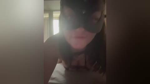 Video of a woman in a black cat mask, lying on a bed, with a blurred, dimly lit room in the background.
