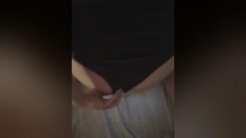 A blurred video showing a person's hands adjusting the waistband of light-colored pants, likely female, wearing a black top. The background is dark, with a dimly lit room setting.