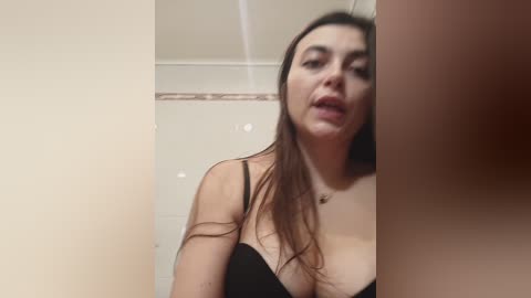 Media: A video of a woman with long brown hair, wearing a black bra, standing in a bathroom with beige walls and a tiled shower.