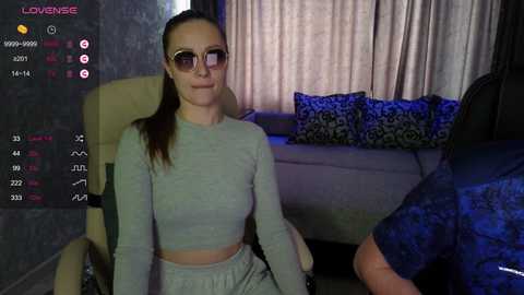 A video of a light-skinned woman with long brown hair and sunglasses, wearing a gray crop top and pants, sitting on a beige couch in a dimly lit room.