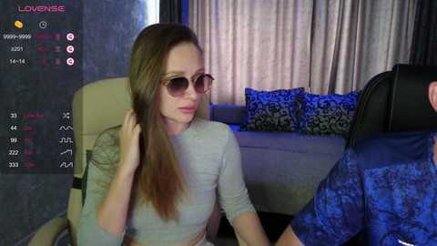 Video of a light-skinned woman with long brown hair, wearing sunglasses and a light gray top, sitting on a couch with blue-patterned pillows, in a dimly-lit room with purple curtains.