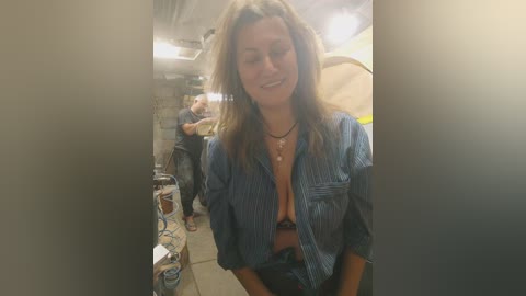 Media: A candid video of a smiling, light-skinned woman with shoulder-length, wavy, blonde hair, wearing a blue striped shirt and a black bra, standing in a dimly lit, cluttered room with a water bottle on a table.