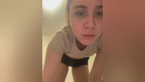 Media: Video of a young woman with fair skin, brown hair, and blue eyes, wearing a grey t-shirt and black shorts, leaning forward with a serious expression.