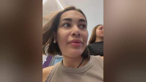 Media: Video of a young woman with medium skin tone, shoulder-length dark hair, and a neutral expression, wearing a light gray ribbed top, seen from the chest up in a modern indoor setting with other people in the background.