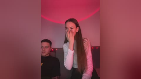 Media: Video of a young woman with long brown hair, wearing a white shirt, covering her face with her hand. A young man in a black shirt sits beside her in a dimly lit room with red lighting.