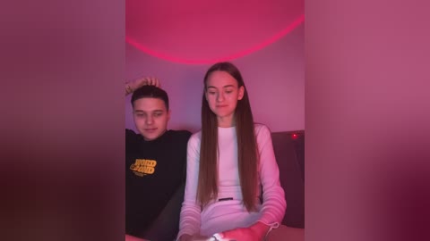 Media: Video of a young couple seated in a dimly lit room with red lighting. The boy, wearing a black shirt, sits behind the girl, who is dressed in white, focusing on a device.