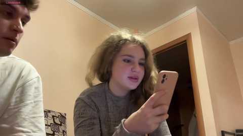 Media: Video of a young woman with curly hair, wearing a gray sweater, taking a selfie in a beige-walled room with a wooden door and patterned art piece.