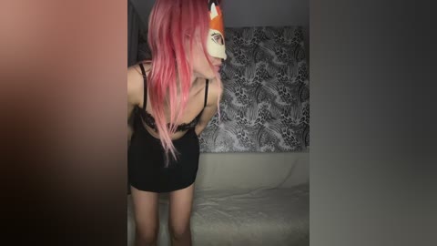 Video of a slender, fair-skinned woman with long, pink hair, wearing a black lace bra and mini skirt, and a white and orange fox mask, standing in a dimly lit room with gray and white patterned wallpaper.