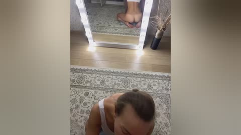 Media: Video of a woman with light skin and short brown hair, wearing a white tank top, lying on a patterned rug in a room with wooden flooring and a lit vanity mirror reflecting her bare feet.