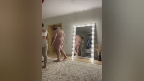 Media: Video of a nude man with a large build and a woman in a white crop top and pants, both reflected in a lit mirror, in a dimly lit room with beige walls.