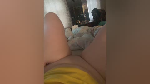 Video of a woman lying on her back, legs spread, wearing yellow shorts, in a messy bedroom with rumpled bedding, a mirror, and a cluttered dresser in the background.