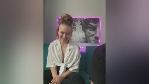 Video of a smiling young woman with light skin and a bun, wearing a white shirt tied at the waist, seated on a teal couch. Background features a black and white portrait and neon purple lighting.