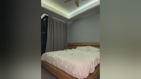 Media: Video of a minimalist bedroom with a wooden bed, white linens, gray curtains, and a ceiling fan. The room has a neutral, serene atmosphere with a modern design.